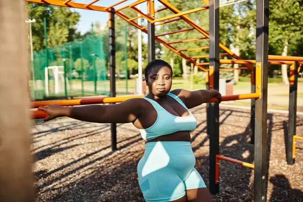 Una mujer afroamericana en ropa deportiva toma una pose frente a un parque infantil, mostrando positividad corporal. - foto de stock