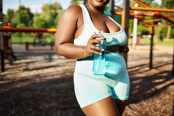 Una mujer afroamericana en un traje de cuerpo blanco, mostrando positividad corporal, sosteniendo una botella de agua mientras hace ejercicio al aire libre. - foto de stock
