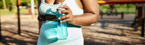 Mujer afroamericana curvilínea en ropa deportiva sosteniendo una botella, rodeada de naturaleza, encarnando la positividad corporal mientras hace ejercicio en un parque. - foto de stock