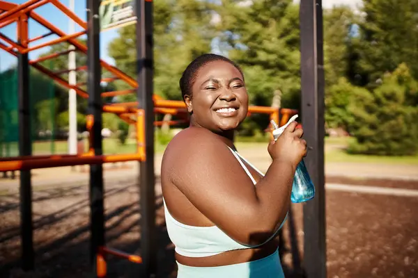 Mujer afroamericana en ropa deportiva se para en el patio de recreo sosteniendo una botella de agua. - foto de stock