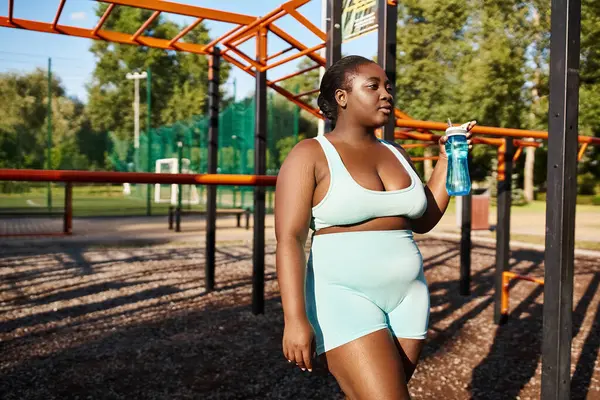 Una mujer afroamericana en ropa deportiva se para en un parque, sosteniendo una botella de agua, tomando un descanso refrescante de su entrenamiento al aire libre. - foto de stock