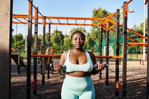 A curvy African American woman in sportswear strikes a powerful pose outdoors, lifts a dumbbells — Stock Photo