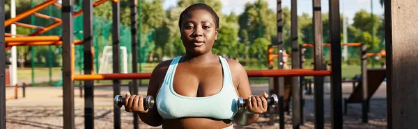 Uma mulher afro-americana com um corpo curvilíneo está trabalhando em um sutiã esportivo azul, segurando halteres na frente de um playground. — Fotografia de Stock