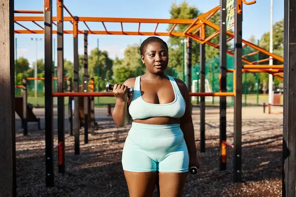 Uma mulher afro-americana curvilínea em um sutiã esportivo azul e shorts azuis claros fica confiante ao ar livre. — Fotografia de Stock