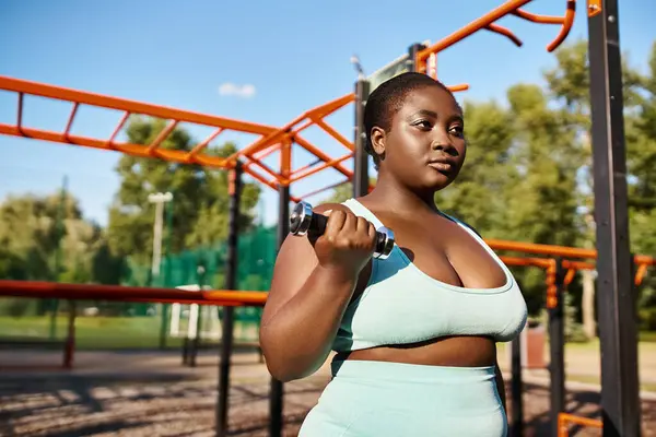 Curvy African American woman in blue top lifting a dumbbell outdoors, embodying strength and confidence. — Stock Photo