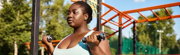 Uma mulher afro-americana em sportswear alegremente levanta halteres na frente de um playground colorido. — Fotografia de Stock