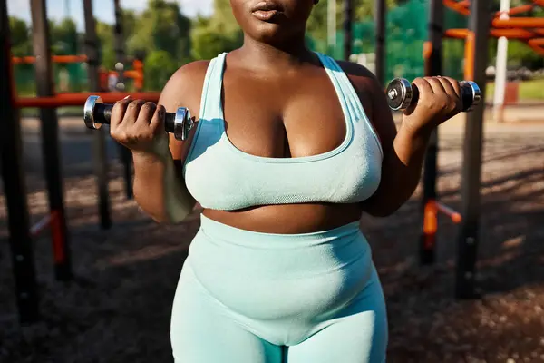 Eine körperlich positive Afroamerikanerin im blauen Sportanzug hebt selbstbewusst zwei Hanteln im Freien. — Stockfoto