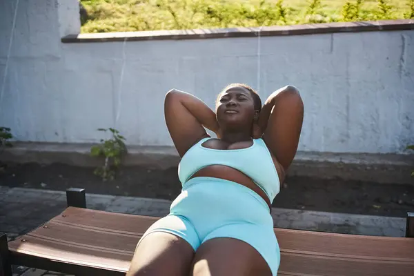 A curvy African American woman in sportswear is laying gracefully on top of a wooden bench outdoors. — Stock Photo