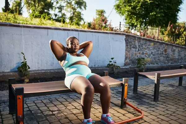 Une afro-américaine courbée, vêtue de vêtements de sport, s'assoit sur un banc les mains derrière la tête, profitant d'un moment de détente. — Photo de stock