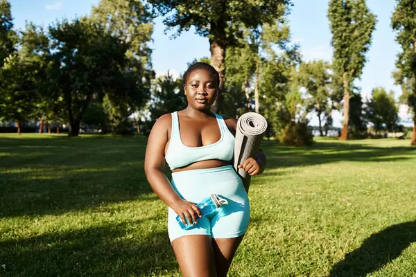 An African American woman in sportswear holding sports mat outdoors, embodying body positivity and strength. — Stock Photo