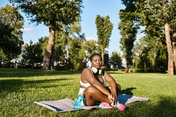 Una mujer afroamericana en ropa deportiva se sienta serenamente en una alfombra deportiva en un parque, encarnando positividad corporal y relajación. - foto de stock