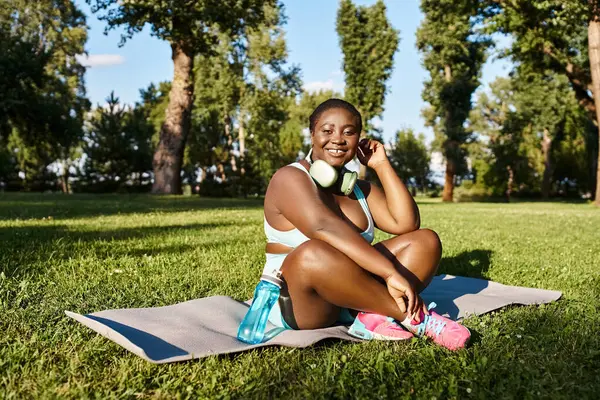 Una mujer afroamericana en ropa deportiva se sienta en una toalla al aire libre, hablando por un teléfono celular. - foto de stock