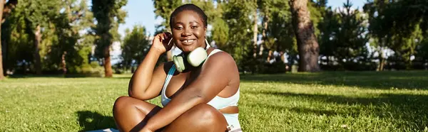 Une afro-américaine courbée en vêtements de sport s'assoit dans l'herbe, appréciant la musique — Photo de stock
