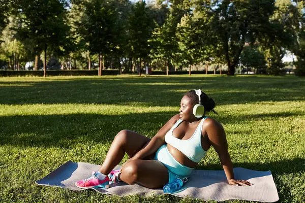 Una mujer afroamericana con curvas en ropa deportiva se sienta pacíficamente en una toalla en un parque, tomando un momento para descansar y admirar la naturaleza. - foto de stock