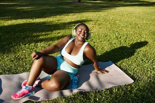 Una mujer afroamericana con curvas en ropa deportiva sentada con gracia en una esterilla de yoga practicando mindfulness al aire libre. - foto de stock