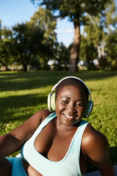 An African American woman in sportswear sits in the grass, listening to music through headphones, enjoying the outdoors. — Stock Photo