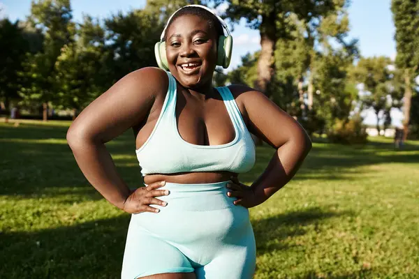 An African American woman in sportswear stands in the grass, embracing body positivity, while listening to music through headphones. — Stock Photo