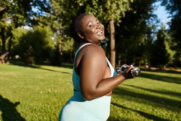 A body-positive African American woman in sportswear holds two dumbbells while exercising in a serene park setting. — Stock Photo