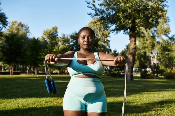 Уверенная афроамериканка в спортивной одежде, поднимающая скакалку в безмятежном парке, демонстрирующая позитивность и силу тела. — стоковое фото