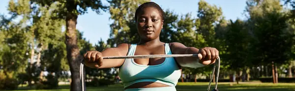 Eine Afroamerikanerin, körperlich positiv und stark, hebt ein Springseil in einer lebendigen Parklandschaft. — Stockfoto
