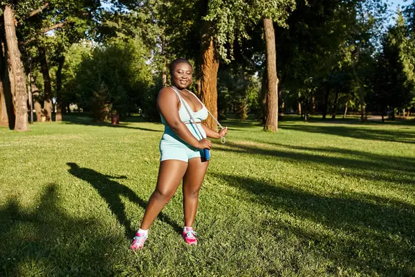 Una mujer afroamericana en ropa deportiva se para en la hierba, sosteniendo con confianza saltar la cuerda - foto de stock