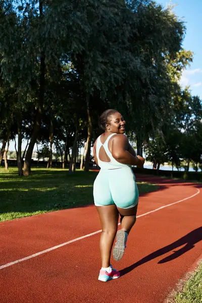 A body-positive African American woman in sportswear is running on a track outdoors, showcasing grace and strength. — Stock Photo