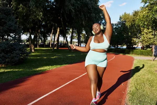 Афроамериканка в спортивной одежде уверенно бежит по красной дорожке под открытым небом, олицетворяя позитивность и силу тела.. — стоковое фото