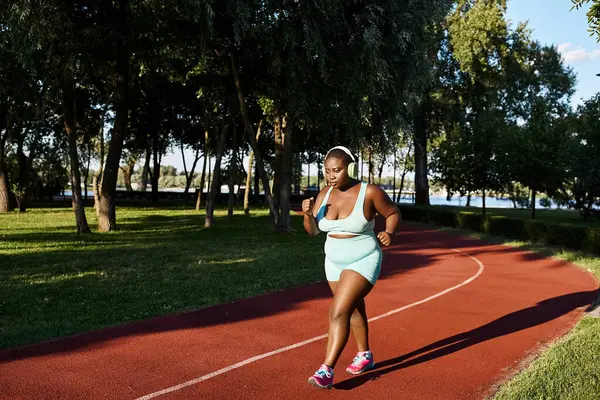 Uma mulher afro-americana curvilínea em sportswear corre energeticamente em uma pista em um parque verde. — Fotografia de Stock