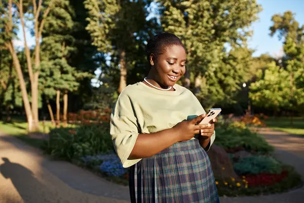 Una mujer afroamericana de talla grande con atuendo casual parada en un parque, absorta en su teléfono celular en un día soleado. - foto de stock