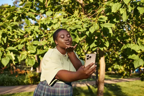 Plus Size Afroamerikanerin steht unter einem Baum und hält an einem sonnigen Tag ein Handy in der Hand. — Stockfoto