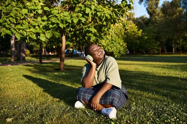 Una mujer afroamericana de talla grande se sienta en la exuberante hierba, absorta en la música bajo el cálido sol de verano. - foto de stock