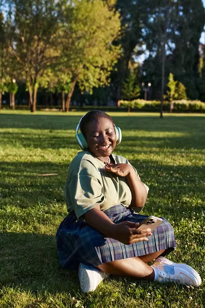 Una mujer afroamericana de talla grande se sienta en la hierba, con auriculares, disfrutando de la música en un día soleado de verano. - foto de stock