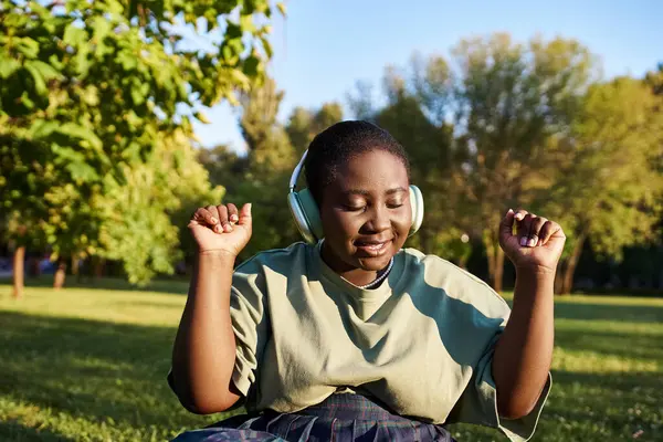 Una mujer afroamericana de talla grande, con atuendo casual, se sienta en la hierba en verano, escuchando música con auriculares. - foto de stock