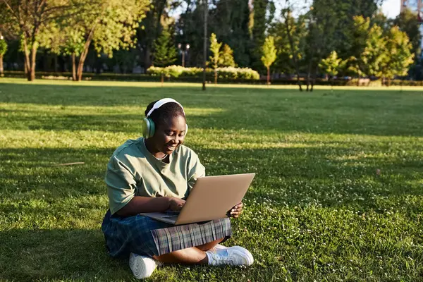 Плюс розмір афроамериканська жінка насолоджується природою під час роботи на ноутбуці в траві в сонячний день. — стокове фото