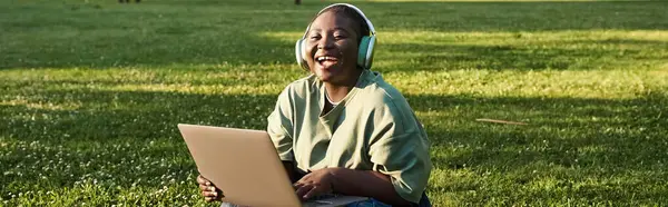 Una mujer afroamericana de talla grande se sienta en un prado cubierto de hierba, absorta en su computadora portátil en un día soleado. — Stock Photo