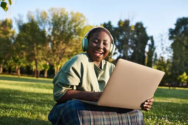 Tamaño más mujer afroamericana sentada en la hierba con el ordenador portátil, disfrutando del trabajo al aire libre en el entorno de verano. — Stock Photo