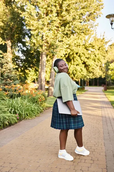 A confident African American woman with a beautiful smile wearing a green sweater and plaid skirt, promoting body positivity outdoors in summer. — Stock Photo