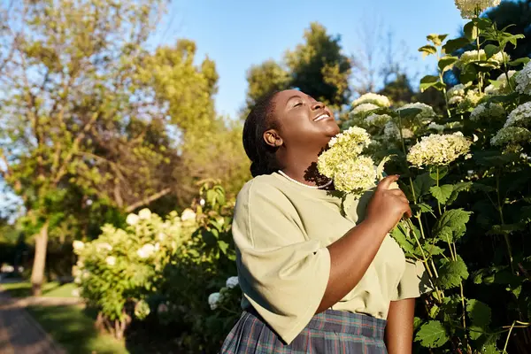 Una mujer afroamericana de talla grande con atuendo casual se para con gracia junto a un arbusto de flores vibrantes al aire libre en el verano. - foto de stock