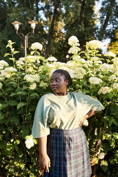Une femme afro-américaine de grande taille se tient en confiance devant un buisson aux fleurs blanches, embrassant la beauté de la nature autour d'elle. — Photo de stock