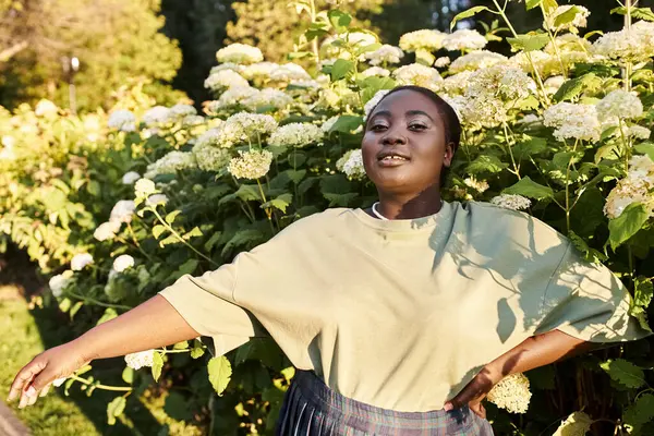 Une femme afro-américaine de taille plus se tient en confiance devant un buisson de fleurs vibrantes, incarnant la positivité du corps et embrassant la beauté de la nature. — Photo de stock