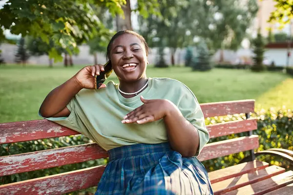 Mujer afroamericana de talla grande con atuendo casual, sentada en un banco al aire libre en el verano, hablando por teléfono celular. - foto de stock