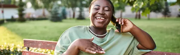 Una mujer afroamericana de talla grande con atuendo casual se sienta en un banco al aire libre, charlando animadamente en su teléfono celular. - foto de stock