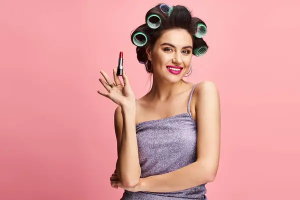 Stylish woman with curlers in hair holding lipstick in front of vibrant backdrop. — Stock Photo