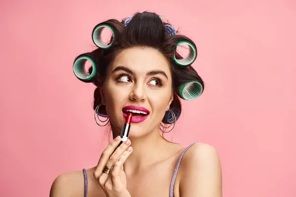 A stylish woman with curlers in her hair using lipstick against a vibrant backdrop. — Stock Photo