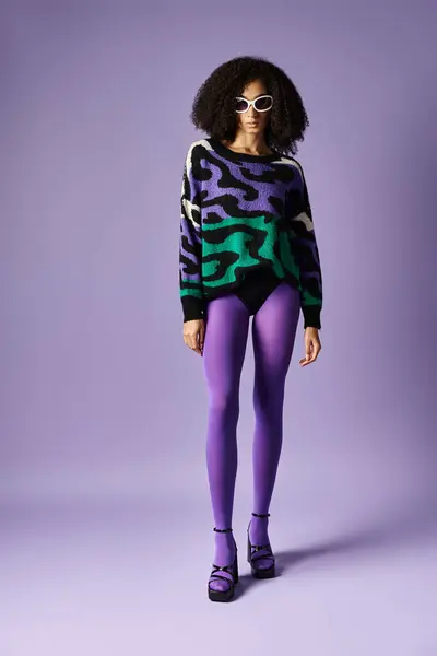 A young woman striking a pose in a vibrant purple and green sweater and purple tights against a purple studio backdrop. — Stock Photo
