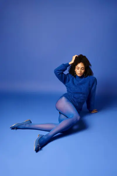 Young woman sitting gracefully on the floor, legs crossed, wearing vibrant tights and sweater against a blue background. — Stock Photo