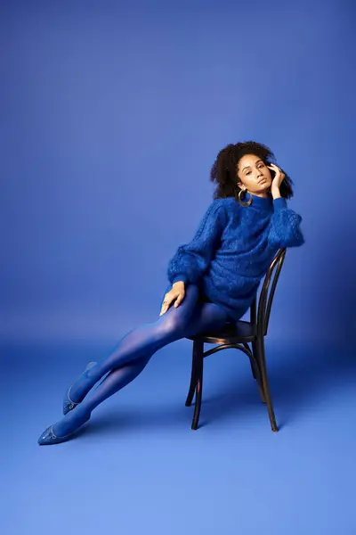Una joven con un elegante vestido azul se sienta elegantemente en una silla sobre un vibrante fondo de estudio azul. - foto de stock