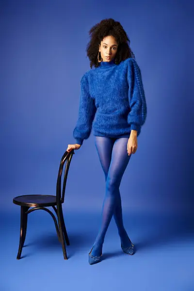 Giovane donna in calzamaglia vibrante e maglione colpisce una posa su una sedia contro uno sfondo blu in un ambiente di studio. — Foto stock