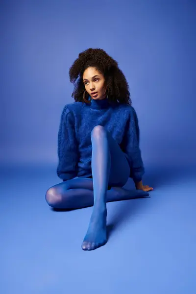 Uma jovem e elegante em meias vibrantes e suéter fica de pernas cruzadas em um chão de estúdio azul, exalando serenidade e postura. — Fotografia de Stock