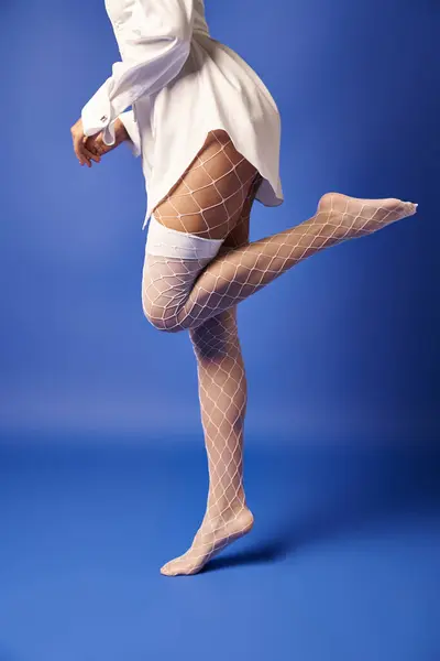 Una mujer joven en medias blancas y vestido golpea una pose elegante en un estudio con un fondo azul vivo. - foto de stock